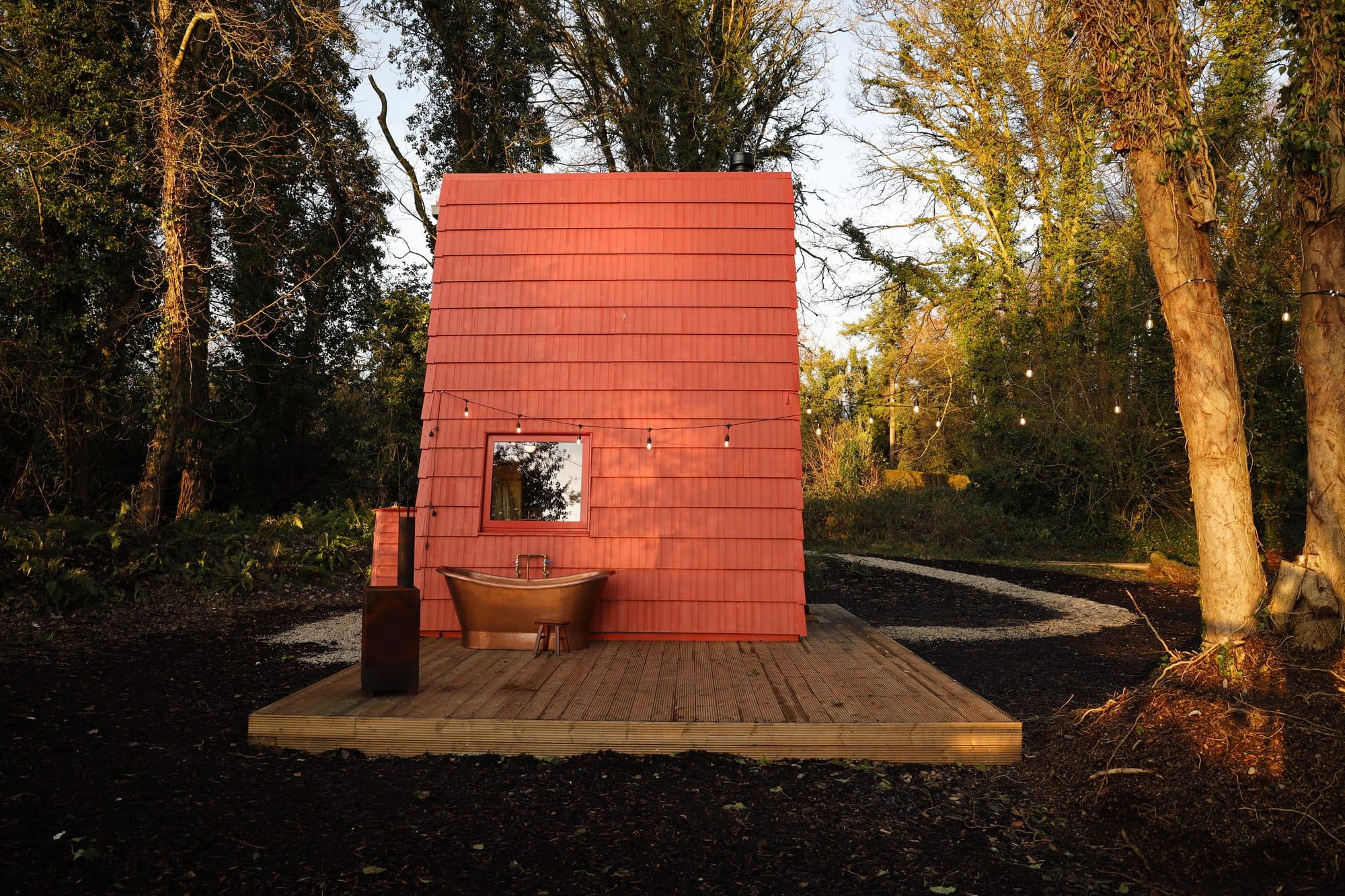 Cucu, a tall red-shingled cabin on a timber deck in a woodland clearing, with a copper bath outside.