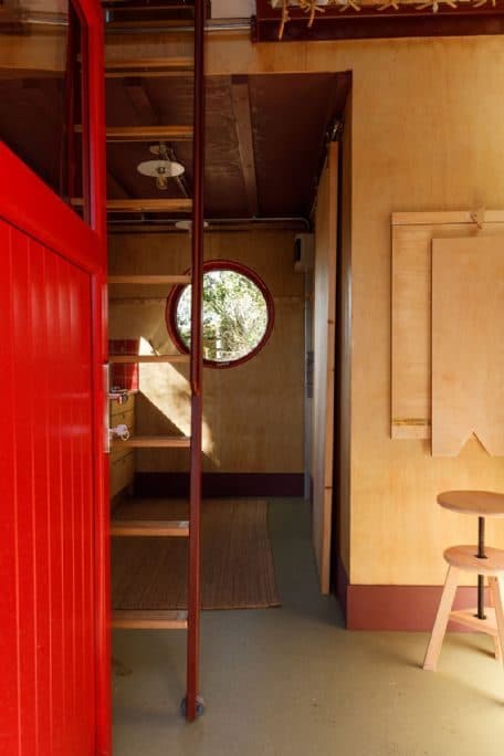 Inside Cucu: plywood walls, a red ladder, a porthole window and a small wooden stool.