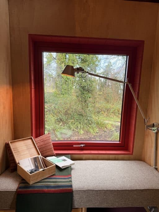 A red-framed picture window over a cushioned reading nook, looking out onto woodland.