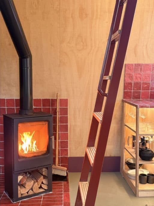A black wood-burning stove on a red-tiled hearth, with the ladder to the mezzanine beside it.