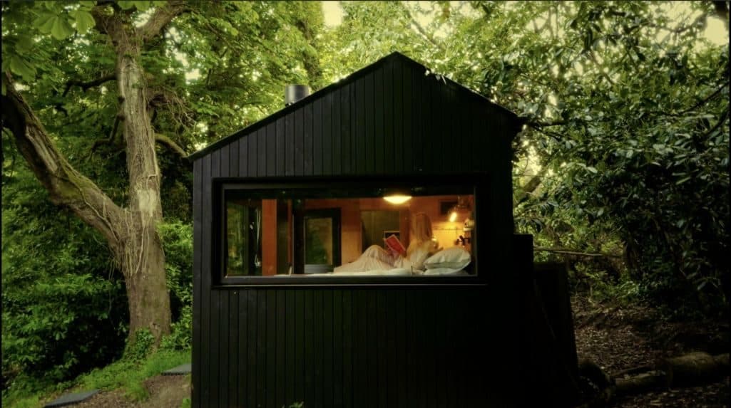 Saoirse at dusk: a black timber cabin among the trees, its long window lit warmly from inside.