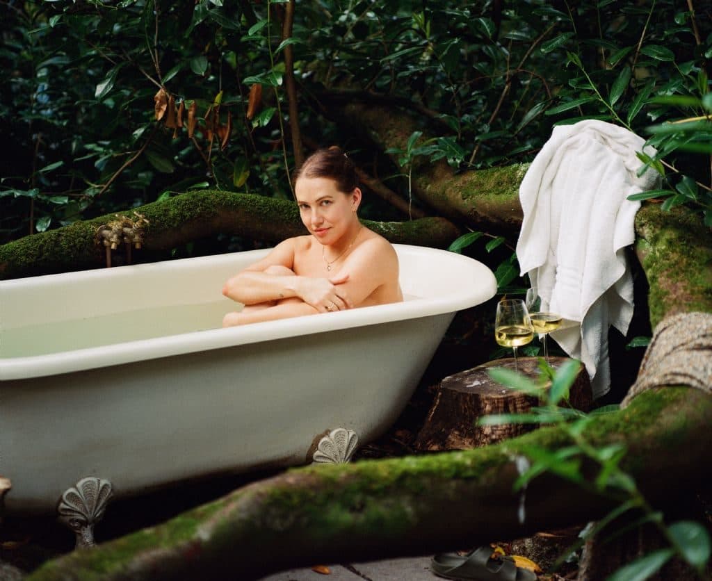 A roll-top bath set among mossy trees, with a towel hung on a branch beside it.