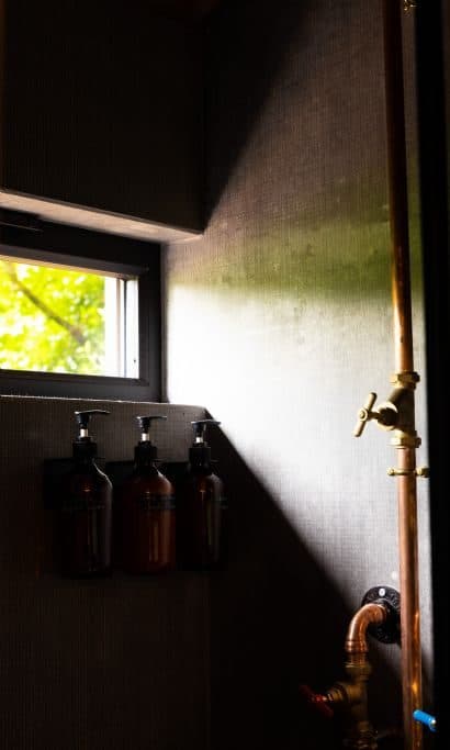 Organic toiletries beside the copper pipework of the shower, lit by a small window.